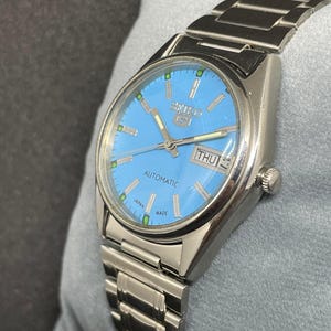 Pu&ograve; includere: Un orologio automatico Seiko 5 color argento con quadrante azzurro. L'orologio presenta un display giorno-data e un cinturino in acciaio inossidabile. Il quadrante ha indicatori verdi e la scritta "SEIKO 5 AUTOMATIC JAPAN MADE".