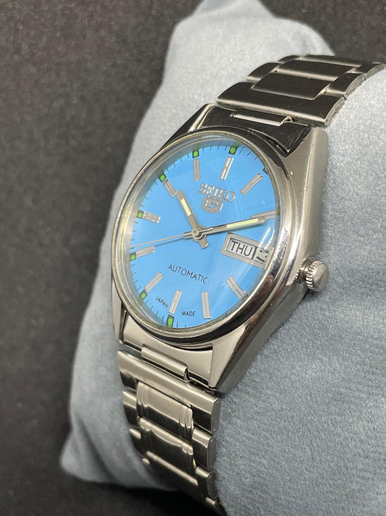 Pu&ograve; includere: Un orologio automatico Seiko 5 color argento con quadrante azzurro. L'orologio presenta un display giorno-data e un cinturino in acciaio inossidabile. Il quadrante ha indicatori verdi e la scritta "SEIKO 5 AUTOMATIC JAPAN MADE".