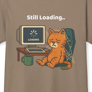 May include: A beige t-shirt featuring a cartoon illustration of a dejected orange cat slumped at a desk with a computer. The screen shows a loading symbol. Text above reads "Still Loading..". A green mug is on the desk.