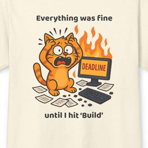 May include: A cream-coloured t-shirt with a distressed orange cat illustration, surrounded by flames and papers. The text reads "Everything was fine until I hit 'Build'" with a computer screen displaying "DEADLINE". A humorous design.
