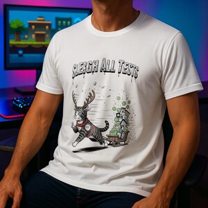 Op de afbeelding: Wit t-shirt met de tekst "SLEIGH ALL TESTS" boven een cartoonkat met geweien en een harnas, die een slee vol cadeaus trekt. De kat is grijs en wit met zwarte strepen. De slee is bruin.