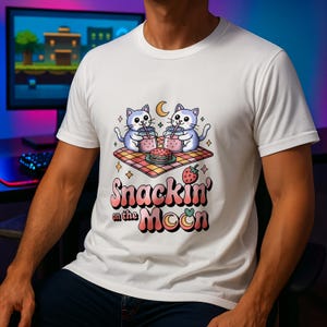 May include: White t-shirt with a cartoon illustration of two cats enjoying pink drinks, a burger, and a strawberry on a picnic blanket. The text "Snackin' on the Moon" is below the image in a playful font.