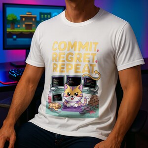 Puede incluir: Camiseta blanca con el texto "COMMIT. REGRET. REPEAT." en letras doradas. Debajo, un gato de dibujos animados en un ordenador con tres monitores. El gato es morado y amarillo. La camiseta es de cuello redondo.