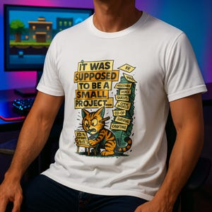 Könnte beinhalten: Weißes T-Shirt mit einer humorvollen Grafik. Das Design zeigt eine Cartoon-Katze mit einem Bleistift und den Text "IT WAS SUPPOSED TO BE A SMALL PROJECT..." zusammen mit spielbezogenen Begriffen wie "VR", "Quests" und "Crafting".
