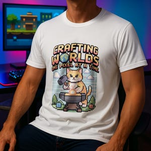 Può includere: T-shirt bianca con grafica pixelata di un gatto che forgia una sfera luminosa su un'incudine, con un martello. Il testo "Crafting Worlds One Pixel At A Time" è sopra il gatto. Tema: videogioco.