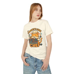 A woman wearing a white t-shirt with a graphic design of a cat wearing a chef's hat and holding a chef's hat.