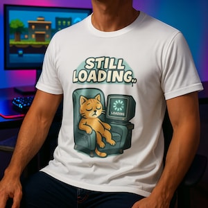 Op de afbeelding: Wit t-shirt met een cartoonafbeelding van een slapende oranje kat in een teal fauteuil, naast een retro computermonitor met een laadsymbool. De tekst "STILL LOADING.." staat boven de kat.