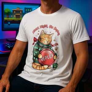 May include: White t-shirt with a cartoon ginger cat wearing a headset and Christmas jumper, holding a red "DEPLOY" button. The shirt has the text "Silent Night, No Deploy" and a "NOEL" banner. A mug of coffee is next to the cat.