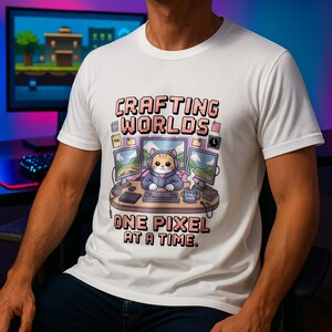 Op de afbeelding: Wit t-shirt met een kleurrijke afbeelding van een kat met een koptelefoon en een hoodie, zittend aan een bureau met computermonitoren. De tekst luidt "Crafting Worlds One Pixel at a Time."