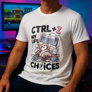 Könnte beinhalten: Weißes T-Shirt mit dem Aufdruck "CTRL + Z MY LIFE CHOICES". Das Design zeigt eine Comic-Katze, die in bunten Kabeln, einer Tastatur und einem Gamecontroller gefangen ist. Ein lässiges Kleidungsstück.