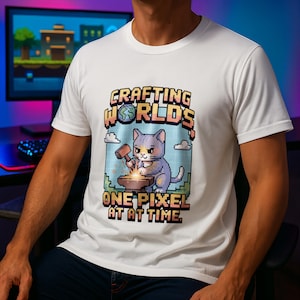 Op de afbeelding: Wit t-shirt met een pixelachtige afbeelding van een grijze kat die op een aambeeld hamert. De tekst luidt "Crafting Worlds One Pixel at a Time" in een retro lettertype. Het ontwerp bevat een gepixeleerde wereld en wolken.