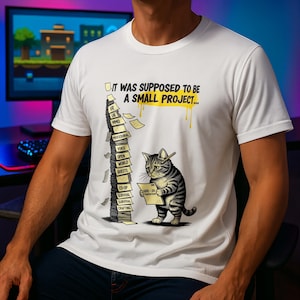 Könnte beinhalten: Weißes T-Shirt mit einer Grafik einer Katze, die ein Papier und einen Stapel Papiere mit Begriffen der Spieleentwicklung hält. Der Text auf dem Shirt lautet: "IT WAS SUPPOSED TO BE A SMALL PROJECT..."