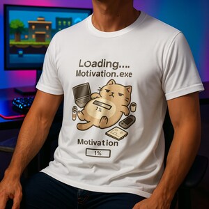 Op de afbeelding: Wit T-shirt met een cartoon kat ontwerp en de tekst "Loading.... Motivation.exe" en "Motivation 1%". De kat is omringd door gaming accessoires. Het shirt is gemaakt van een zacht, comfortabel materiaal.