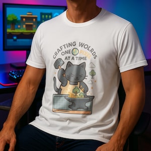 Peut inclure: T-shirt blanc avec un chat forgeron gris tenant un marteau au-dessus d'une enclume. Le chat porte un tablier jaune et fabrique des objets pixélisés. Le texte dit "Crafting Worlds One Pixel at a Time".