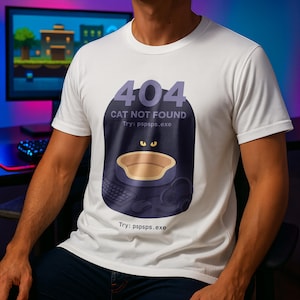 Può includere: T-shirt bianca a maniche corte con grafica. Il design presenta un messaggio di errore 404 a tema gatto, tra cui una cuccia, una tastiera e il testo "CAT NOT FOUND" e "Try: pspsps.exe".