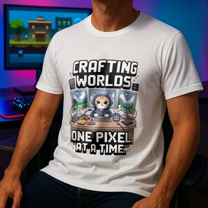 Op de afbeelding: Wit t-shirt met een pixelachtig ontwerp met een kat met een koptelefoon aan een computerbureau. De tekst luidt "Crafting Worlds One Pixel at a Time."
