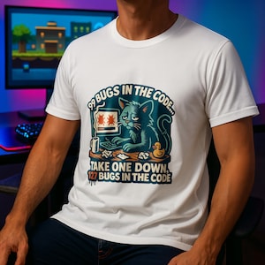Op de afbeelding: Wit T-shirt met een vervaagde afbeelding van een blauwe kat die op een computer werkt. De tekst luidt: "99 BUGS IN THE CODE... TAKE ONE DOWN, 127 BUGS IN THE CODE." Een badeend en een mok zijn ook afgebeeld. Het T-shirt heeft korte mouwen.
