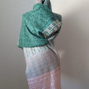 Könnte beinhalten: Ein handgefertigter Schal in den Farben Türkis, Hellblau und Rosa. Der Schal ist über einer Schaufensterpuppe drapiert und zeigt seine komplizierten, spitzenartigen Muster und das farbblockierte Design. Die Kanten haben einen zarten, gewellten Besatz.