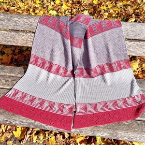 May include: A gray and red knitted scarf with geometric patterns. The scarf features a repeating square design in red along the edges and at intervals throughout the body. The scarf is draped over a wooden bench, with autumn leaves in the background.