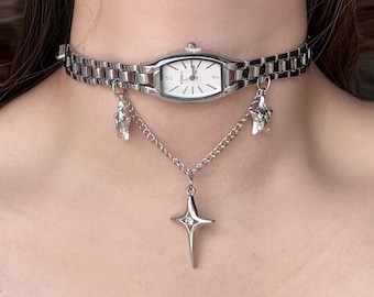 Op de afbeelding: Zilverkleurige choker-ketting met een rechthoekige horlogekast en bungelende ster- en kristallen bedels. Een zilveren ketting met een kruisvormige hanger hangt eronder. De wijzerplaat heeft zwarte wijzers en uurmarkeringen.