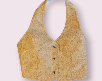 Puede incluir: Un chaleco halter color beige con un patrón floral en relieve. El chaleco tiene tres botones plateados en la parte delantera y un diseño halter. El chaleco está hecho de un material texturizado.