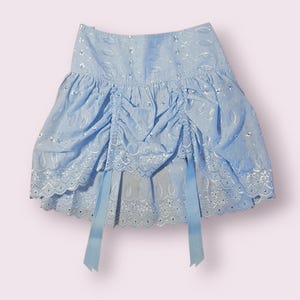 Pode incluir: Uma saia azul clara com cintura franzida e bainha com vieiras. A saia apresenta um delicado padrão floral e é adornada com fitas azuis claras. O tecido tem um brilho subtil, aumentando o seu apelo feminino. Esta saia é uma adição elegante a qualquer guarda-roupa.