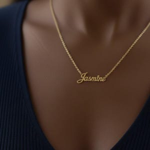 Puede incluir: Un collar dorado con el nombre "Jasmine" en cursiva. El collar se lleva sobre una camiseta azul oscuro. La cadena es delicada y el nombre es el punto focal de la joya.