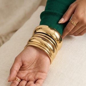 Può includere: Un set di cinque bracciali rigidi in metallo dorato lucido indossati al polso, caratterizzati da una superficie liscia e arrotondata. Il set di gioielli è completato da due anelli a catena dorati abbinati indossati sulle dita, sopra una maglia verde a maniche lunghe.