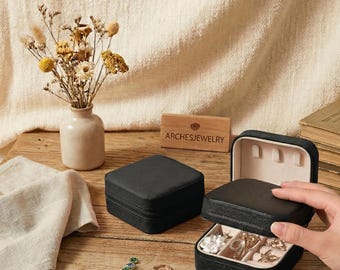 Pode incluir: Estojo de viagem para joias quadrado preto, um aberto para exibir anéis e colares. O estojo está em uma superfície de madeira com outras joias e um pequeno vaso de flores secas. A marca "ARCHESJEWELRY" está em um bloco de madeira.