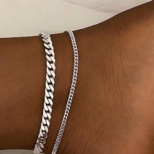 May include: Two silver-toned anklets worn together. One anklet features a thick, flat curb chain design with interlocking links, while the second anklet is a thinner, delicate chain with smaller, tightly woven links, both resting on the skin.