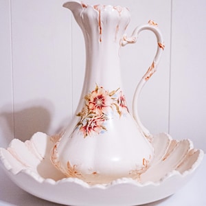 Puede incluir: Una jarra de porcelana blanca vintage con un diseño floral, colocada en una base a juego con bordes festoneados. La jarra presenta un asa curva y está adornada con flores rosas, rojas y azules. Una pieza decorativa clásica.