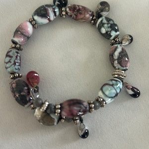 Könnte beinhalten: Armband mit ovalen Perlen in Rosa, Türkis und Schwarz, mit silberfarbenen Akzenten. Die Perlen sind eine Mischung aus opak und durchscheinend, mit tropfenförmigen Anhängern. Einzigartiger Schmuck.