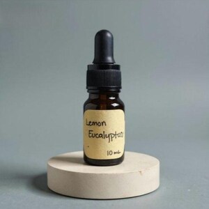 May include: A small amber glass bottle with a black dropper top, labeled "Lemon Eucalyptus" and "10 mL". The bottle sits on a round, light-colored concrete stand against a gray background. The label is a light tan color.
