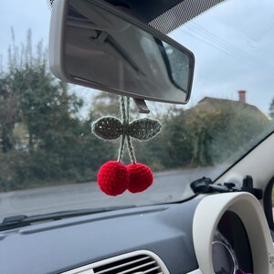 May include: A crochet car charm shaped like two red cherries with green leaves, suspended from a car's rearview mirror. The charm is a decorative item for a vehicle's interior. The car's dashboard and mirror are visible in the background.