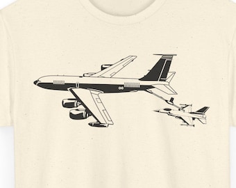 May include: Cream-colored t-shirt featuring a black illustration of a large jet plane and a smaller fighter jet. The graphic is centered on the shirt, offering a minimalist design. The tee is made of a soft fabric.
