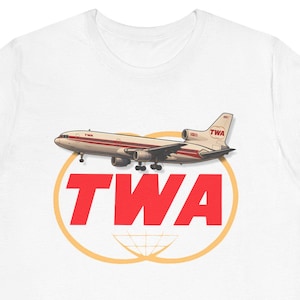 May include: White t-shirt featuring a vintage TWA logo with a red and gold design. The logo includes an airplane illustration above the bold red letters "TWA" and a gold globe outline.
