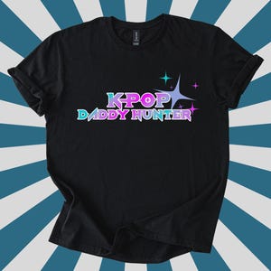 May include: Black t-shirt with the text "KPOP DADDY HUNTER" in a gradient of blue, purple, and pink. The design includes starburst graphics. The shirt is displayed on a blue and white striped background.