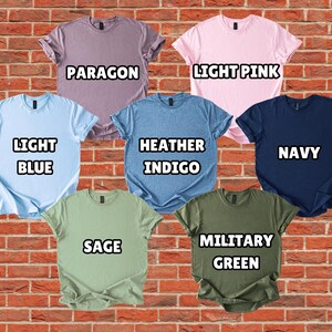 Puede incluir: Una colecci&oacute;n de siete camisetas de manga corta en varios colores, incluyendo azul claro, rosa claro, marino, salvia y verde militar. Cada camiseta tiene un nombre de color diferente impreso en negrita blanca. Las camisetas se muestran sobre un fondo de pared de ladrillo.