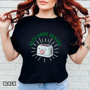 Puede incluir: Camiseta negra con el texto verde "BATH BOMB SEASON", que rodea una tostadora de dibujos animados con un coraz&oacute;n y una cara sonriente. La tostadora es azul claro con detalles blancos.