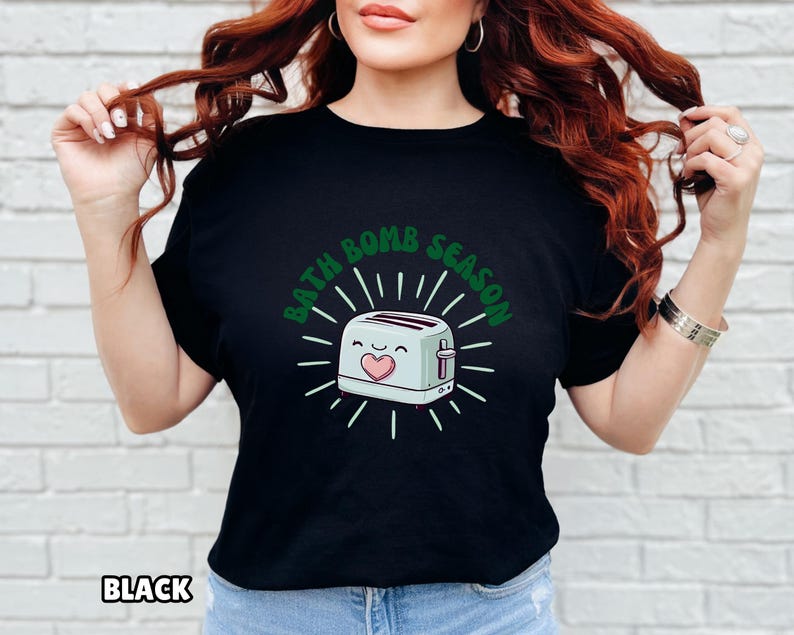 Puede incluir: Camiseta negra con el texto verde "BATH BOMB SEASON", que rodea una tostadora de dibujos animados con un coraz&oacute;n y una cara sonriente. La tostadora es azul claro con detalles blancos.