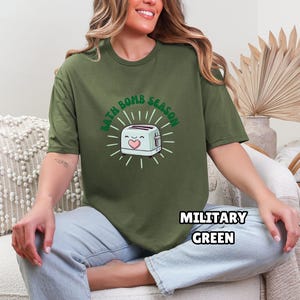Puede incluir: Camiseta verde militar con el texto "BATH BOMB SEASON" que rodea una tostadora de dibujos animados con un coraz&oacute;n. La tostadora es blanca con un coraz&oacute;n rosa y un acento verde. La camiseta es de cuello redondo y manga corta.