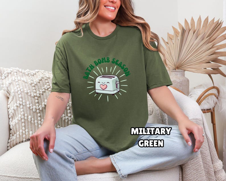 Puede incluir: Camiseta verde militar con el texto "BATH BOMB SEASON" que rodea una tostadora de dibujos animados con un coraz&oacute;n. La tostadora es blanca con un coraz&oacute;n rosa y un acento verde. La camiseta es de cuello redondo y manga corta.