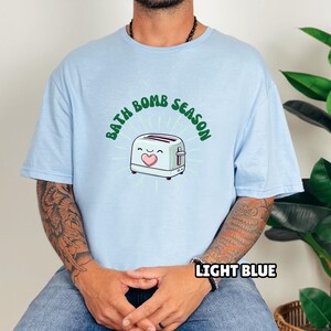 Puede incluir: Camiseta azul claro con el texto "BATH BOMB SEASON" sobre una tostadora de dibujos animados con un coraz&oacute;n. La camiseta tambi&eacute;n tiene el texto "LIGHT BLUE" en la parte inferior. La camiseta es de color azul claro.