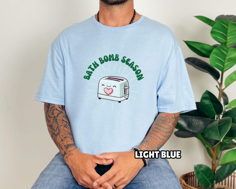 Puede incluir: Camiseta azul claro con el texto "BATH BOMB SEASON" sobre una tostadora de dibujos animados con un coraz&oacute;n. La camiseta tambi&eacute;n tiene el texto "LIGHT BLUE" en la parte inferior. La camiseta es de color azul claro.