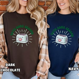 Puede incluir: Dos camisetas, una marr&oacute;n oscuro y otra azul marino, cada una con un dise&ntilde;o de tostadora de dibujos animados y el texto "BATH BOMB SEASON" en verde. La camiseta marr&oacute;n oscuro tiene el texto "DARK CHOCOLATE" en la parte inferior, y la camiseta azul marino tiene el texto "NAVY".