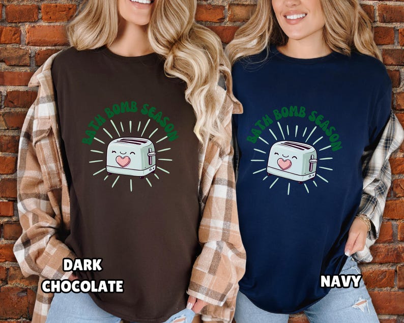 Puede incluir: Dos camisetas, una marr&oacute;n oscuro y otra azul marino, cada una con un dise&ntilde;o de tostadora de dibujos animados y el texto "BATH BOMB SEASON" en verde. La camiseta marr&oacute;n oscuro tiene el texto "DARK CHOCOLATE" en la parte inferior, y la camiseta azul marino tiene el texto "NAVY".