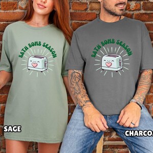 Puede incluir: Camisetas verde salvia y gris carb&oacute;n con el texto "BATH BOMB SEASON" y un dibujo de una tostadora. La tostadora tiene una cara sonriente y un coraz&oacute;n. Las camisetas son de corte holgado.