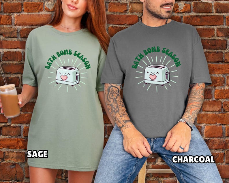 Puede incluir: Camisetas verde salvia y gris carb&oacute;n con el texto "BATH BOMB SEASON" y un dibujo de una tostadora. La tostadora tiene una cara sonriente y un coraz&oacute;n. Las camisetas son de corte holgado.