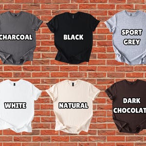 Puede incluir: Se muestran seis camisetas de manga corta en varios colores contra una pared de ladrillos. Los colores incluyen carb&oacute;n, negro, gris deportivo, blanco, natural y chocolate oscuro. Cada camisa tiene una etiqueta con el nombre de su color.