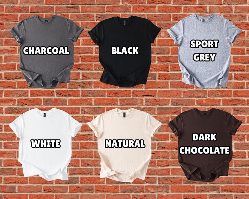 Puede incluir: Se muestran seis camisetas de manga corta en varios colores contra una pared de ladrillos. Los colores incluyen carb&oacute;n, negro, gris deportivo, blanco, natural y chocolate oscuro. Cada camisa tiene una etiqueta con el nombre de su color.